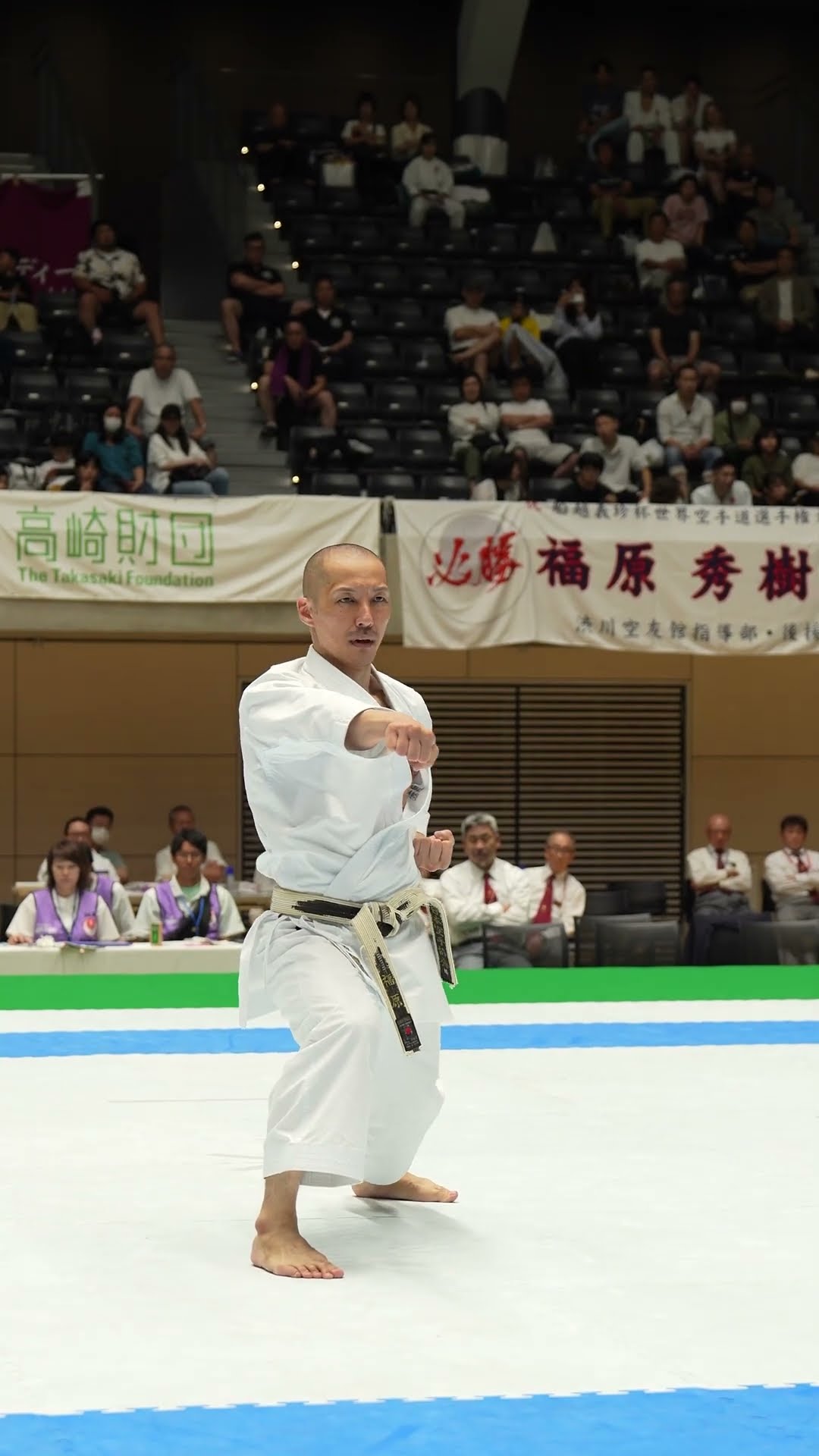 Photo (video still): a JKA karateka in white gi and black belt in a stance with arms raised mid-technique, performing Bassai Sho at a JKA tournament arena