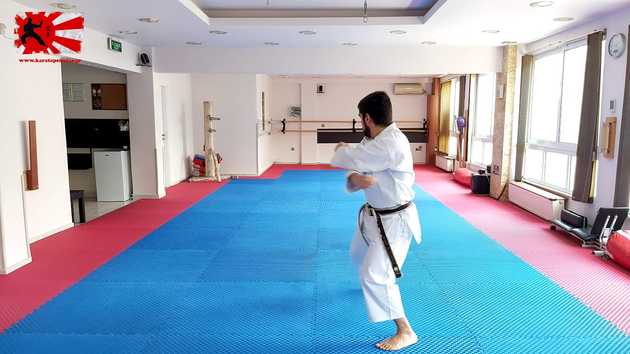 Photo (video still): JKA karateka in white gi and black belt, mid-technique with arms crossed at the chest, performing Wankan on a blue mat in the Peiraias club dojo