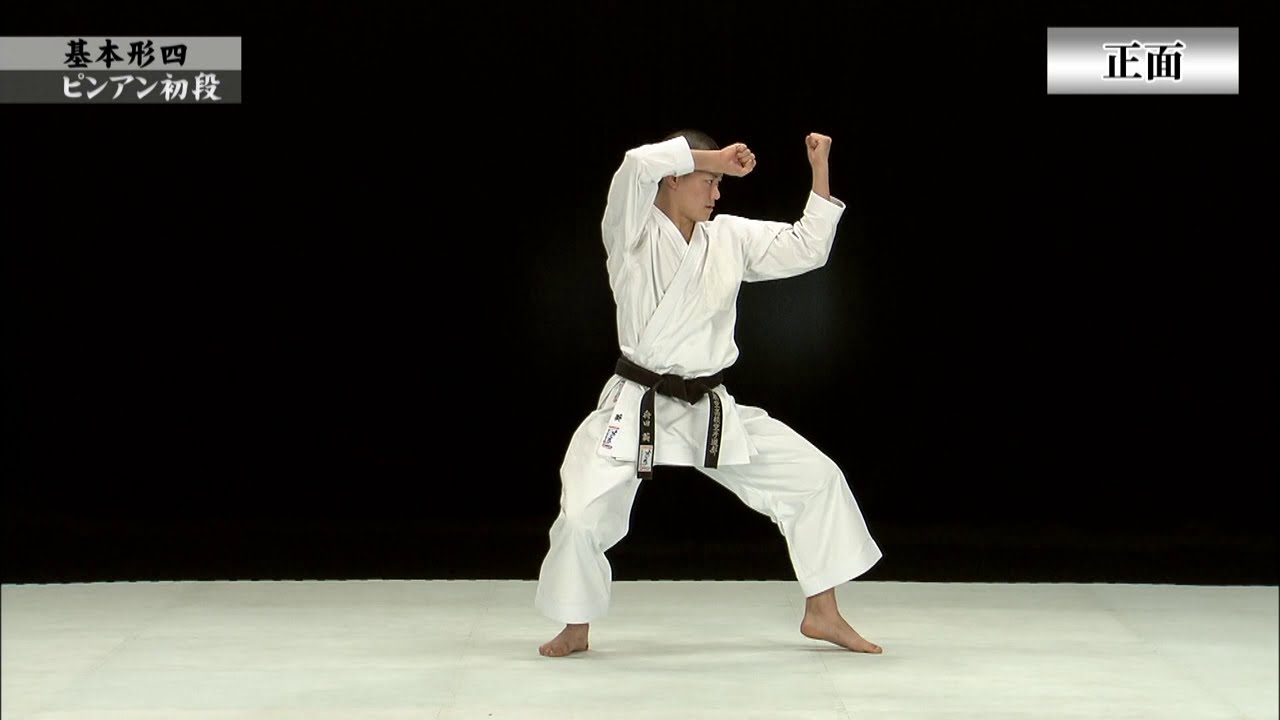 Photo (video still): a JKF demonstrator in white gi and black belt performing Pinan Shodan (Wado-ryu (JKF designated version)), shown full-length mid-technique on a white mat against a black backdrop, with small corner labels
